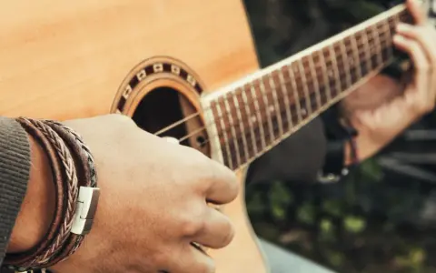 Dłoń z bransoletką z plecionej skóry gra na gitarze akustycznej.