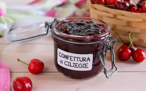 Un vasetto di marmellata di ciliegie fatta in casa, con ciliegie fresche e un cestino sullo sfondo.