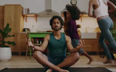 Man i lugn meditation i köket medan barn springer förbi. En lugn stund för 5 minute meditation mitt i vardagskaoset.