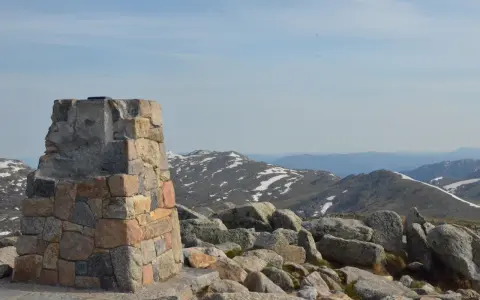 Kamienny monument na szczycie góry Kościuszko, otoczony głazami, z widokiem na ośnieżone pasma górskie w oddali.