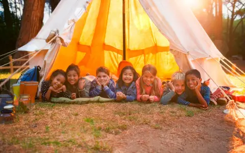 Sept enfants souriants allongés devant une tente de camping éclairée par le soleil.