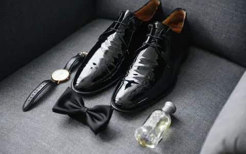 Shiny black patent leather shoes to wear with tuxedo, a bow tie, watch, and cologne are laid out on a grey couch.