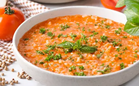 Eine cremige Tomatensuppe mit Reis, garniert mit frischer Petersilie und Basilikum. Daneben liegen frische Tomaten und Gewürzkörner.