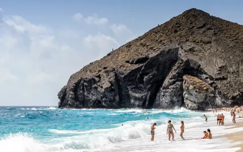 Sonniger Almería Urlaub: Menschen genießen den Strand und das Meer vor einer imposanten Felsformation.