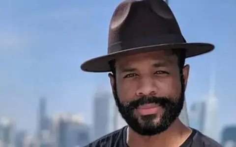 Hombre con sombrero y barba espesa, sonriendo ante un fondo de edificios y cielo azul.