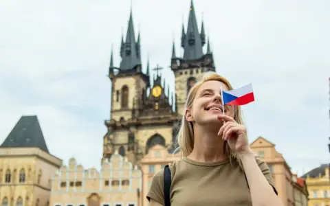 Uśmiechnięta blondynka z flagą Czech zasłaniającą oko, marzy o wysokich zarobkach w Czechach. W tle praskie kamienice i kościół.