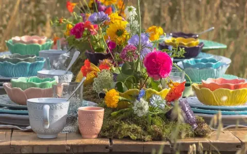 Färgglada uppläggningsfat i plast, staplade med blommor och glas, skapar en festlig dukning utomhus.