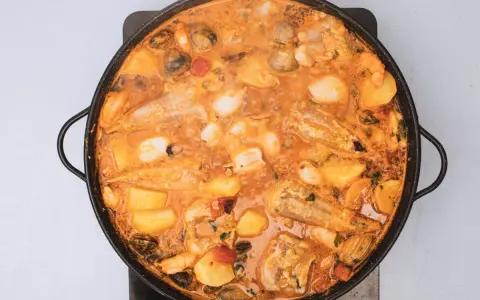 Un suquet de peix burbujeante en una paellera, con trozos de pescado, patatas, gambas y mejillones en una salsa naranja.