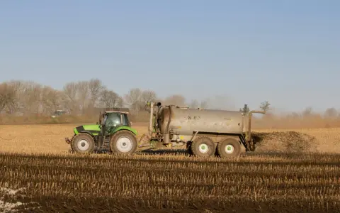 Zielony traktor z przyczepą rozprowadza obornik bydlęcy na polu.