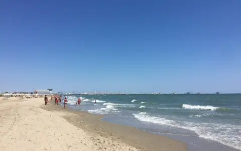 Spiaggia affollata a Ravenna, con gente che passeggia sulla sabbia e nel mare. All'orizzonte, barche e un cielo azzurro intenso.