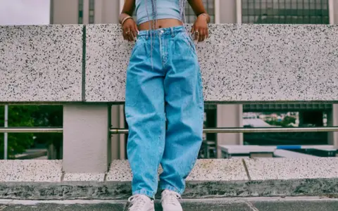 Person in einem lässigen baggy jeans outfit mit Crop-Top und weißen Sneakern steht auf einer Betonmauer.