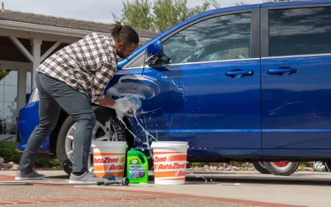 A man washes a blue car in the sun, with buckets and car wash soap nearby. It's a good day to wax a car in the sun.