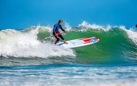 Ein Mann im Neoprenanzug surft auf einem SUP-Board eine grüne Welle. Das SUP-Wellenreiten erfordert Balance und Geschick.