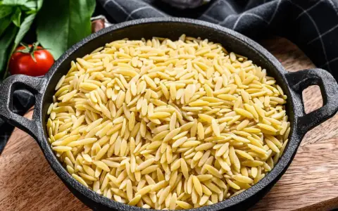 Makaron orzo w czarnej patelni, obok pomidorów i ziół. Wygląda jak ryż, ale to pyszny makaron!