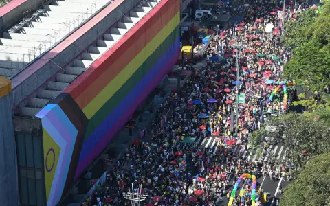 A massive crowd fills the street for what appears to be the biggest pride parade, with a giant rainbow flag draped over a building.