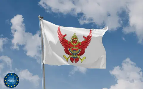 Biała flaga z wizerunkiem czerwonego Garudy, symbolu Tajlandii, powiewa na tle błękitnego nieba z chmurami.