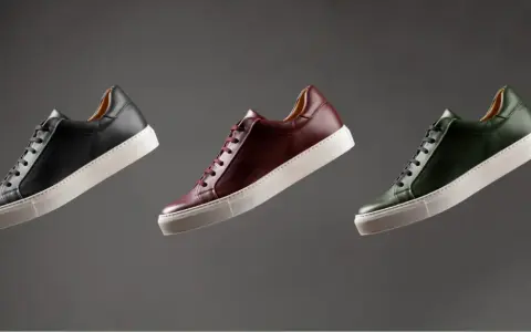 Sneakers marche famose in pelle nera, bordeaux e verde, con suola bianca, fluttuano su sfondo grigio.