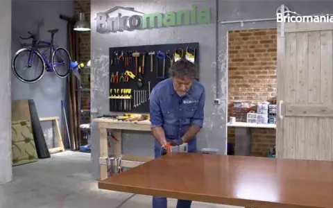 Hombre lijando mesa de madera. El taller tiene herramientas colgadas y una bicicleta morada. La madera hinchada por humedad se repara.