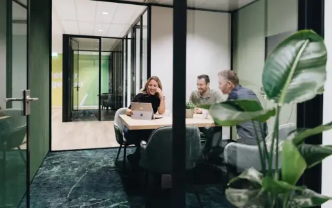 Tre kollegor skrattar vid ett mötesbord i en modern, inredd verkstad. En kvinna visar något på sin laptop.