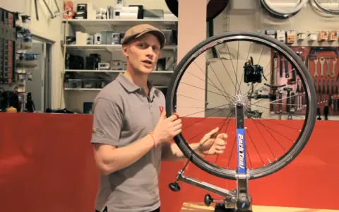 Man i keps visar hur man rikta cykelhjul i en verkstad. Verktyg hänger på väggen bakom.