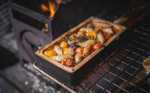 Salsiccia grillen in einer Keramikform mit Tomaten und Kräutern. Flammen lodern im Hintergrund.