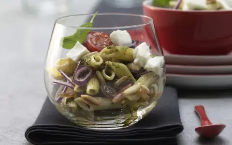 Ein köstlicher Nudel Pesto Salat mit Mozzarella, Pinienkernen, Rosinen und roten Zwiebeln in einem Glas serviert.