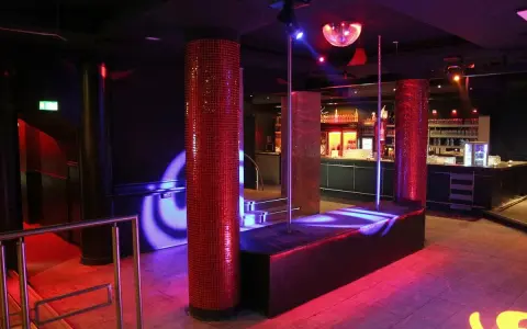 Interno di una boite de nuit con luci soffuse, un palo da lap dance, un bancone bar e una sfera da discoteca.