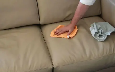 Mano che pulisce un divano in tessuto non sfoderabile con un panno arancione. Accanto, un panno grigio piegato.