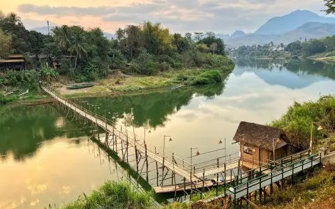 Drewniany mostek nad rzeką, bambusowa chatka i góry w tle. To jedne z wielu **laos atrakcje** warte zobaczenia.