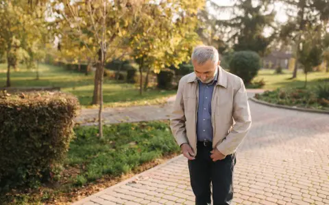 Starszy mężczyzna w beżowej kurtce Harrington i niebieskiej koszuli spaceruje po parku jesienią.