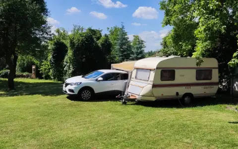 Biały samochód i przyczepa kempingowa na zielonej trawie, otoczone drzewami. Idealne miejsce na **camping smok**.