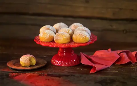 En röd tårtfat med lussebullar med kvarg, pudrade med socker. En ensam bulle ligger på en träplatta.