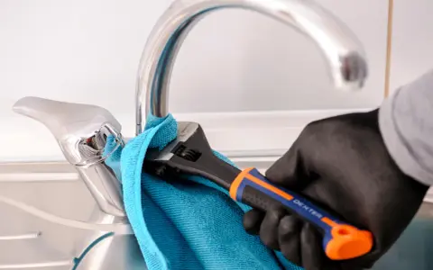 Mano enguantada usa llave inglesa para cambiar grifo cocina. Toalla azul protege el cromo.