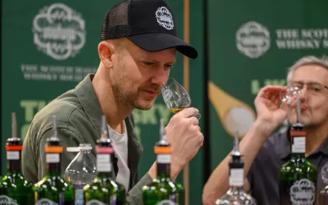 Mann mit Kappe riecht an einem Whiskyglas bei einer Whisky Messe. Flaschen stehen auf dem Tisch.