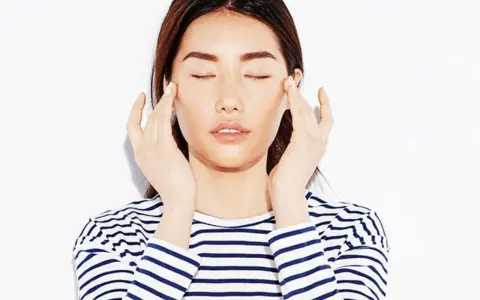 A woman with closed eyes performs a gentle face massage for puffiness, her fingers delicately touching her temples.