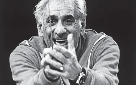 Uśmiechnięty Leonard Bernstein, dyrygent, z dłońmi złożonymi przed sobą, wpatruje się w widza.