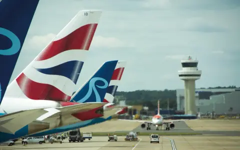 Samoloty z charakterystycznymi ogonami British Airways i innym logo czekają na start na lotnisku Londyn Gatwick. W tle wieża kontroli lotów.