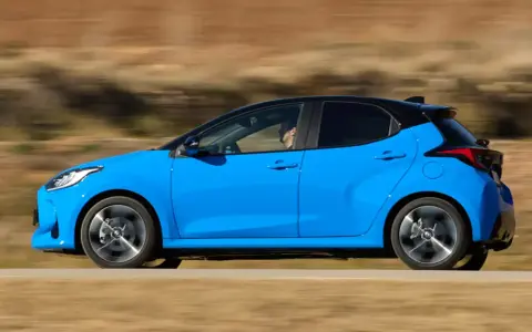 Niebieski hatchback z czarnym dachem pędzi po drodze, za kierownicą siedzi mężczyzna.