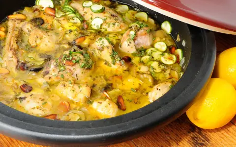 Tajín de pollo con calabacín, aceitunas y almendras, servido en una cazuela de barro, con limones frescos al lado.