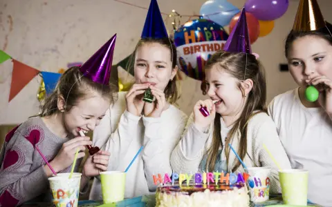 Radosne zabawy na urodziny! Dziewczynki w stożkowych czapkach dmuchają w gwizdki, śmiejąc się przy torcie z napisem "Happy Birthday".