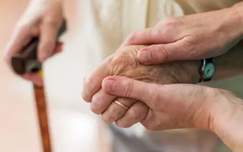 Dwie dłonie, jedna starsza, druga młodsza, trzymają się nawzajem. Laska w tle sugeruje trudności z poruszaniem się, być może związane z chorobą Parkinsona i jej dziedziczeniem.