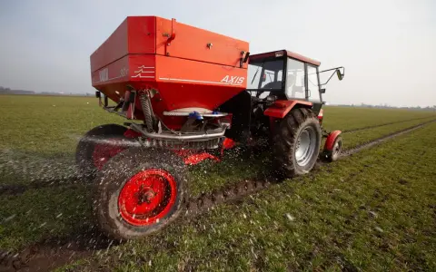 Czerwony traktor z rozsiewaczem nawozu na wiosennym polu. Odpowiedni nawóz pod warzywa na wiosnę to klucz do obfitych plonów.
