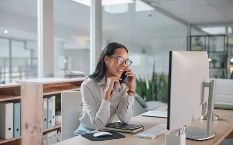 Donna sorridente al telefono in ufficio, mentre lavora al computer. Potrebbe chiedersi quanto guadagna una segretaria al mese.