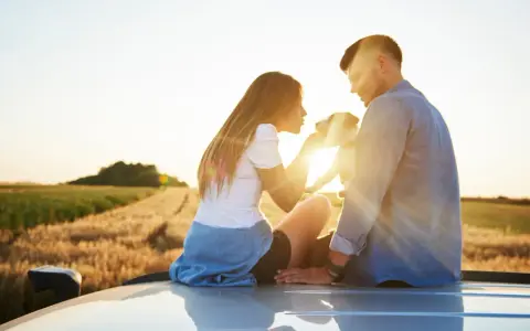 Ein Paar sitzt auf einem Auto im Sonnenuntergang, ein Hund zwischen ihnen. Sie scheinen in einer stillen Partner Meditation zu sein, umgeben von Feldern.