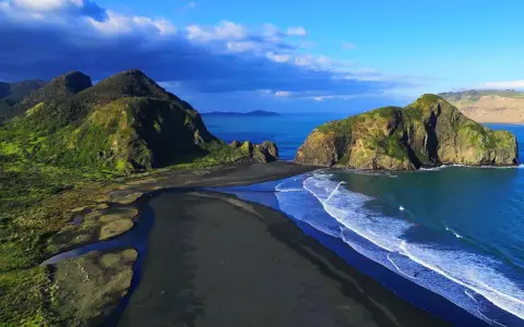 Vacker svart sandstrand med gröna kullar och klippor vid Auckland, Nya Zeeland. Havet skiftar i blått.