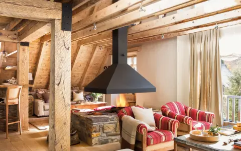 Chimenea central de diseño moderno, uno de los tipos de chimeneas que aporta calidez a este salón rústico con vigas de madera.