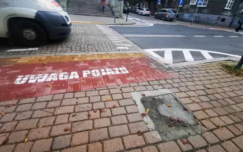 Czerwony pas na chodniku z napisem "UWAGA POJAZDY" informuje o przejściu sugerowanym. W tle widać samochody i schody.