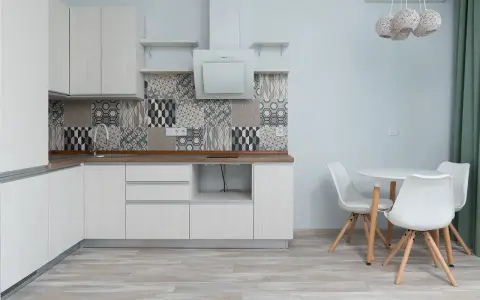 Cocina moderna con armarios blancos, salpicadero geométrico y suelo gris claro. Mesa redonda con sillas blancas.