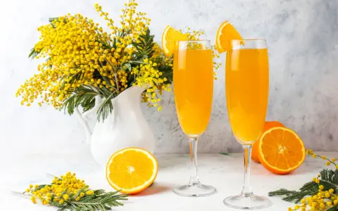 Dwa kieliszki orzeźwiającego drinka mimosa, ozdobione plasterkami pomarańczy, stoją obok bukietu żółtych kwiatów akacji.