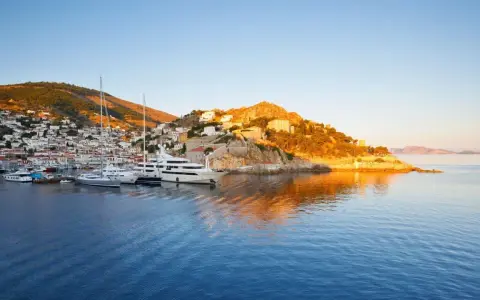 Solnedgång över hamnen på Hydra, Grekland. Lyxyachter guppar i det blå vattnet framför en vit stad som klättrar uppför en grönskande kulle.